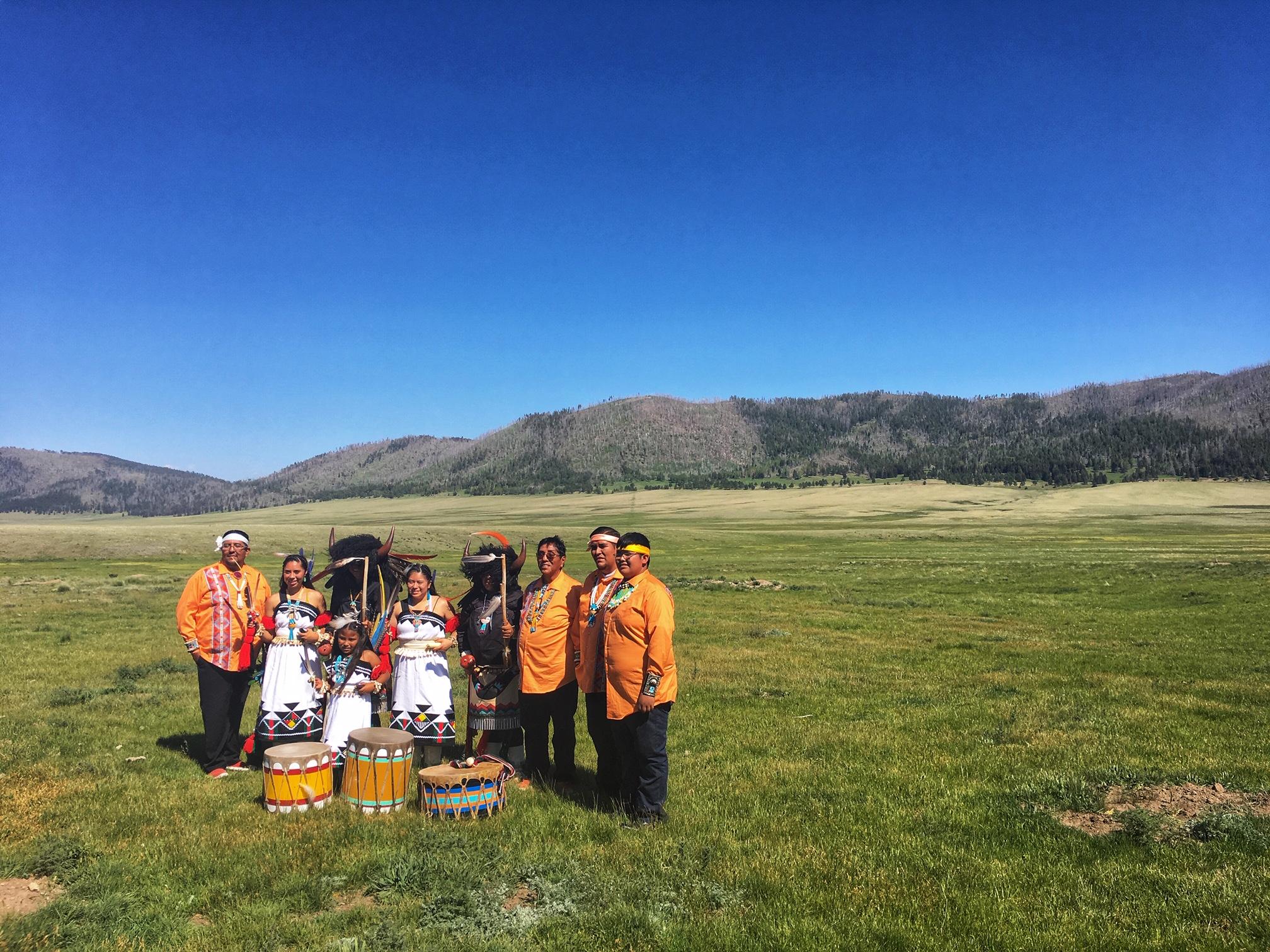A group of indigenous dancers dressed in traditional regalia stands in a large montane grassland.