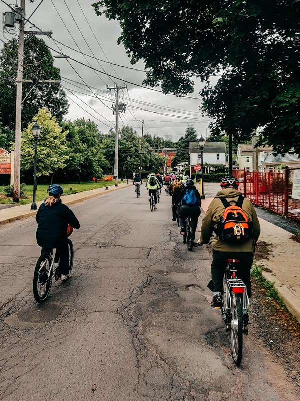 A group of cyclists ride on right shoulder of a city street on a cloudy day