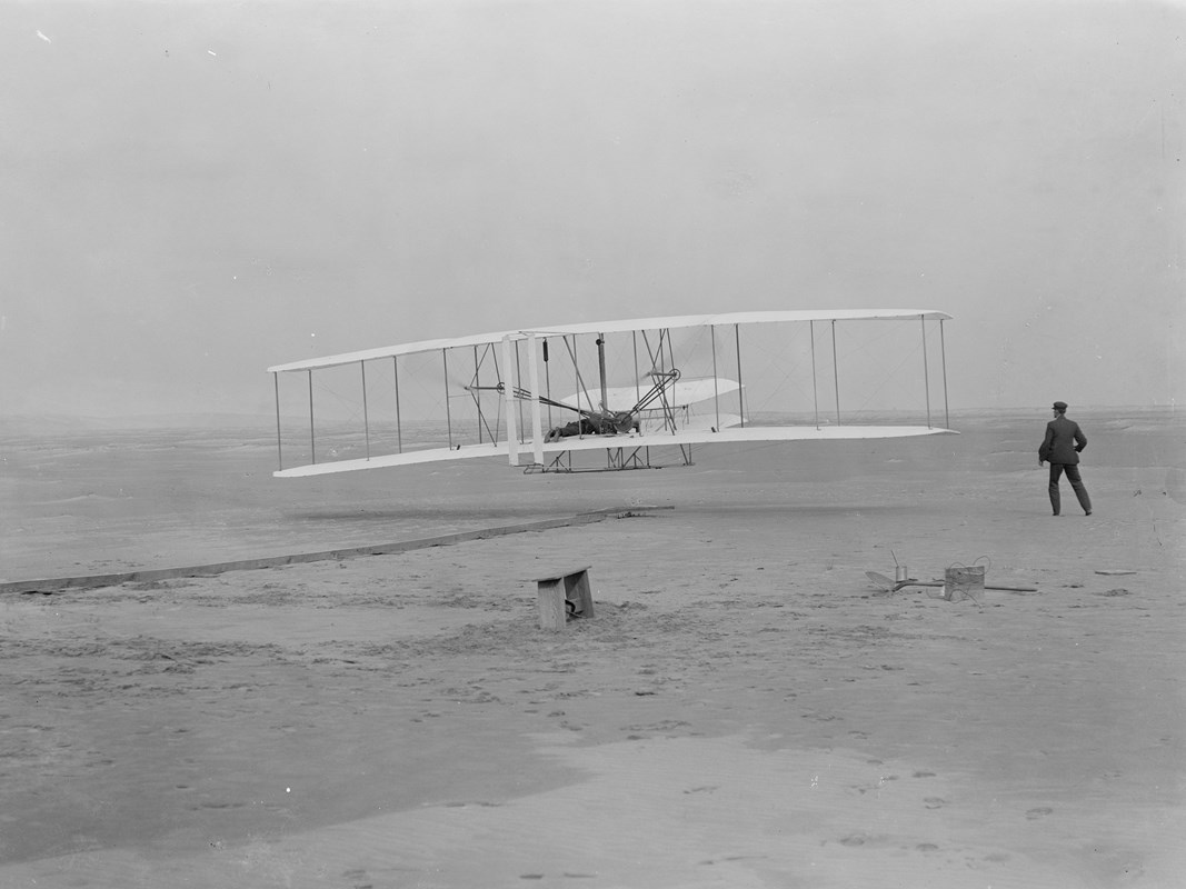 The 1903 Wright Flyer, a small bi-plane, flying a few feet above the sand with Wilbur standing by.