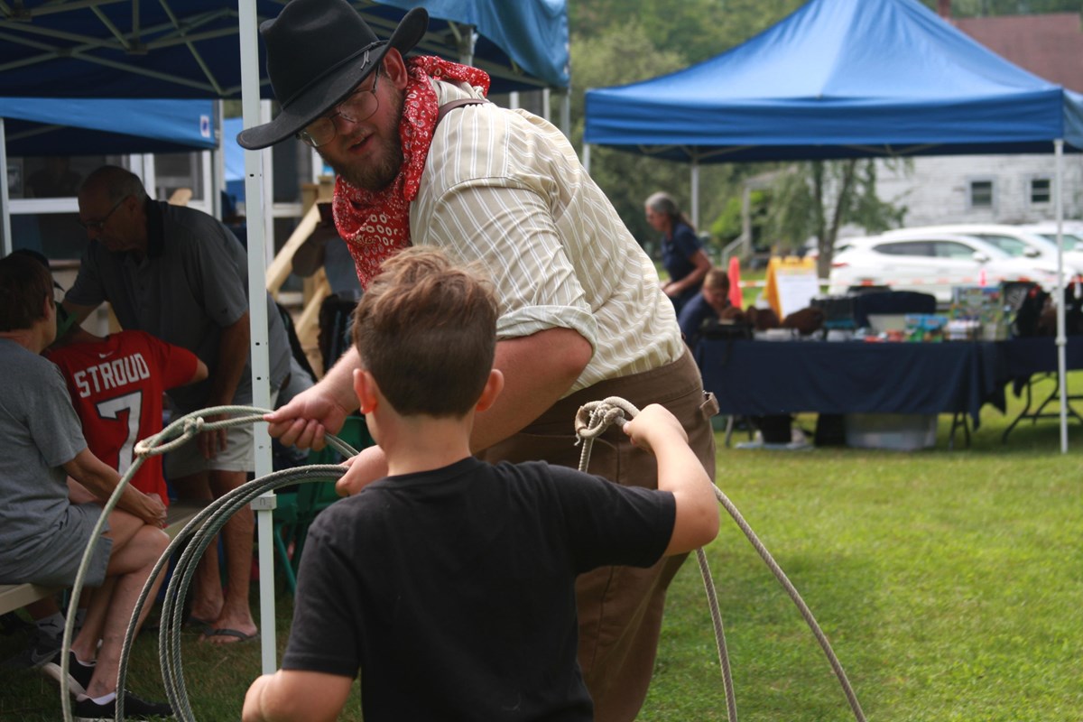 Man in cowboy hat and neckerchief shows young boy how to handle a rope lasso.