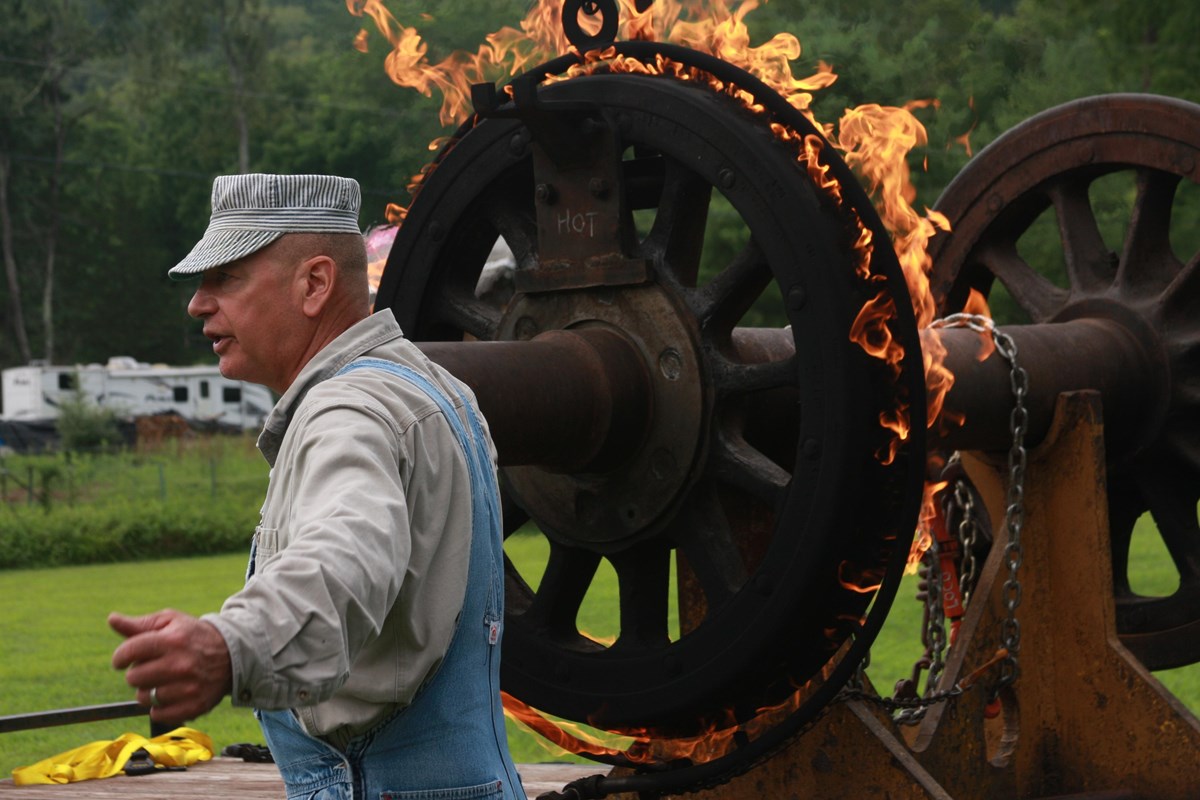 Man in old fashioned train engineer cap and overalls stands in front of large metal on-fire wheel.