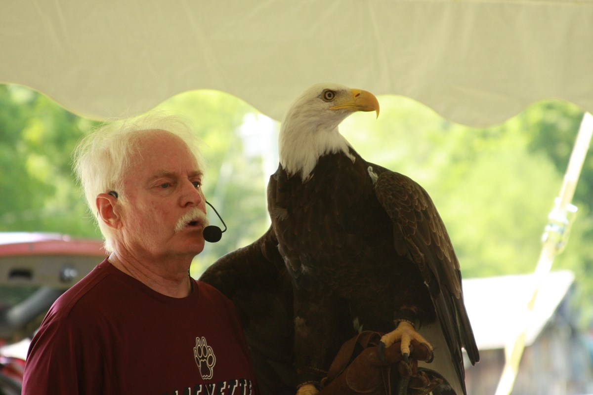 Older man with a microphone and a bald eagle perched on his arm.