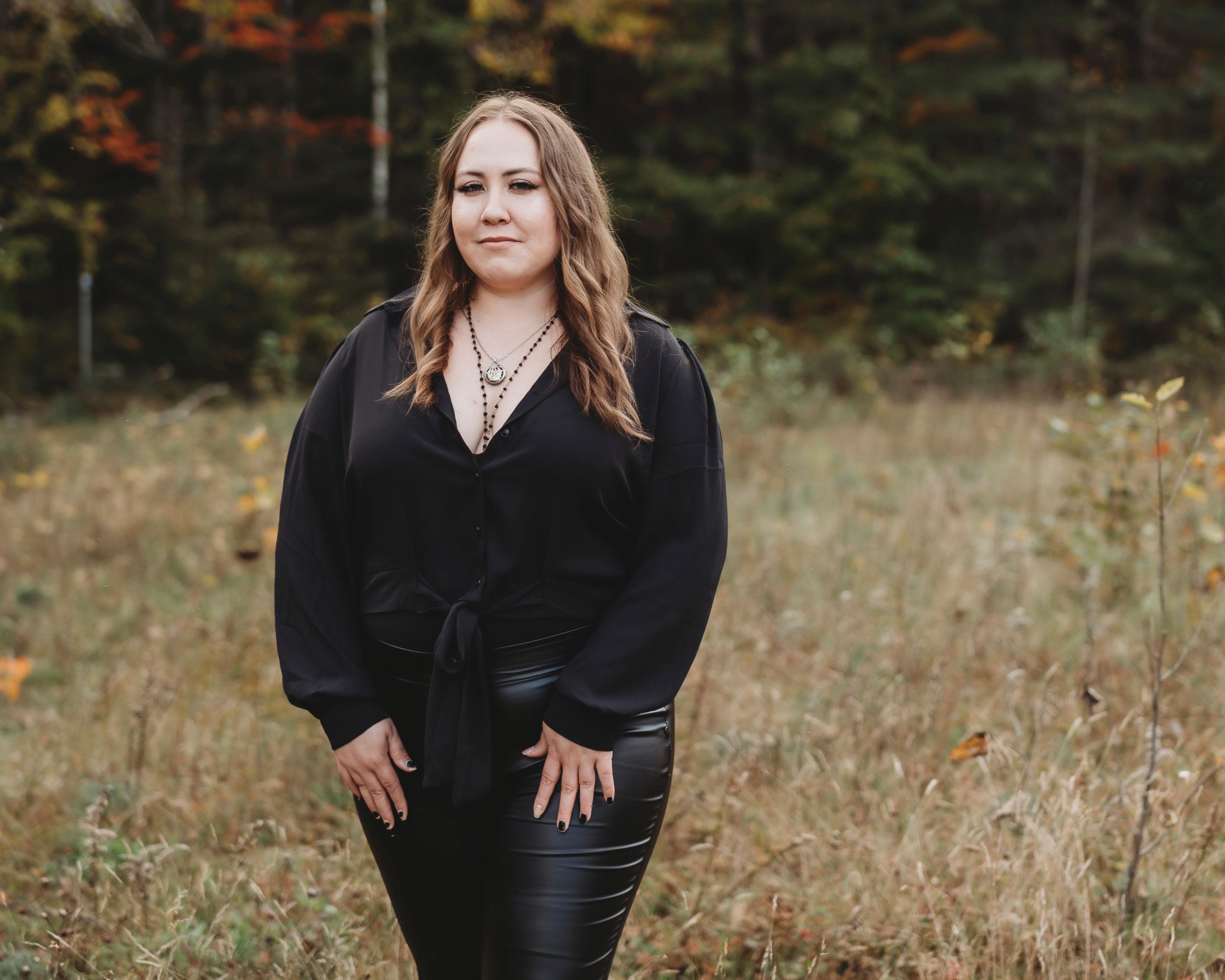 Woman with long wavy brown hair and wearing black stands in a field with trees in distance
