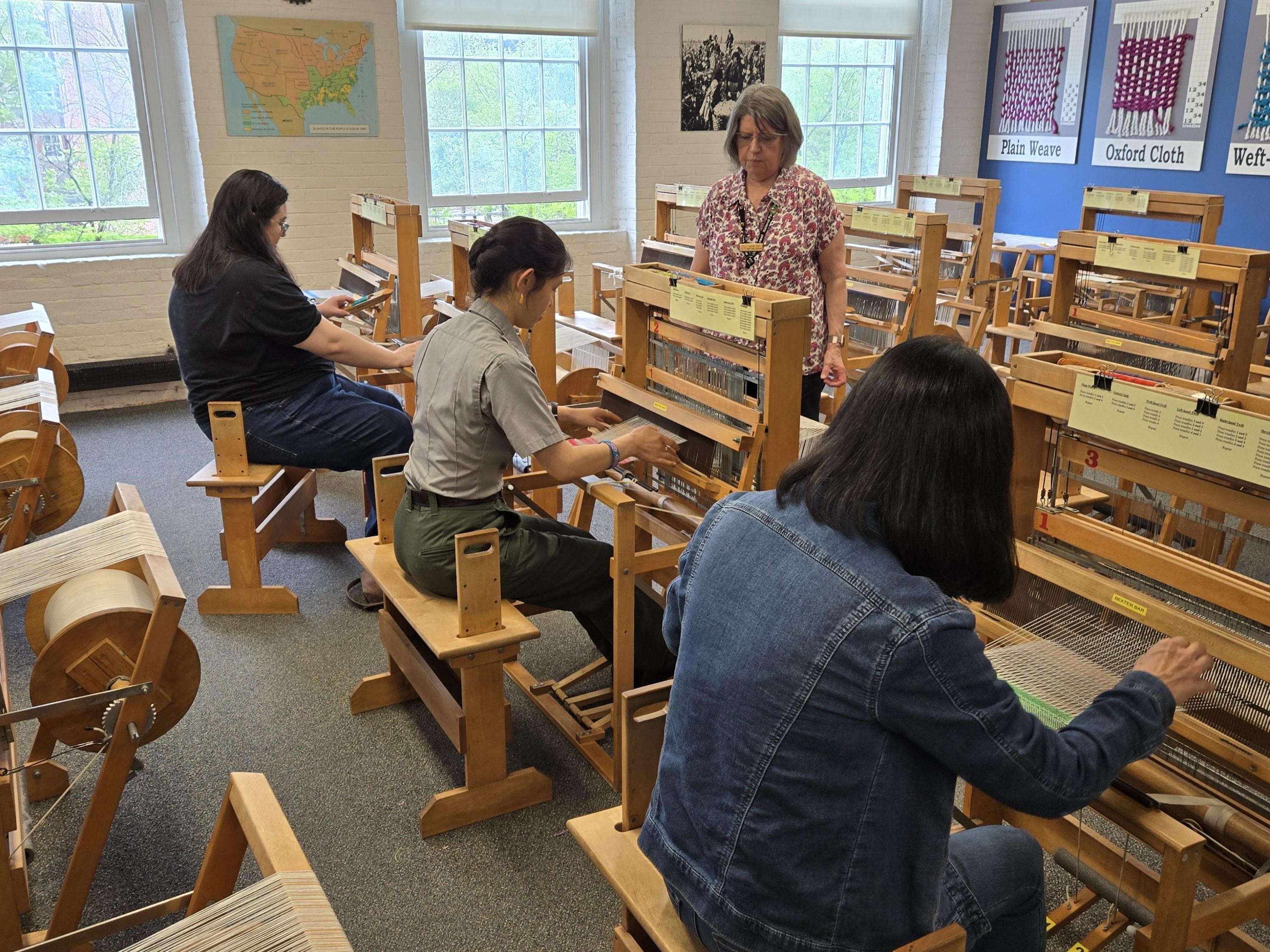 A ranger sits at a hand loom with two visitors on either side, while an instructor walks in front.