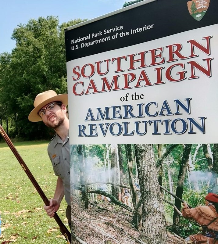 A man in gray shirt and straw ranger hat holding an antique rifle peeks from behind a poster.