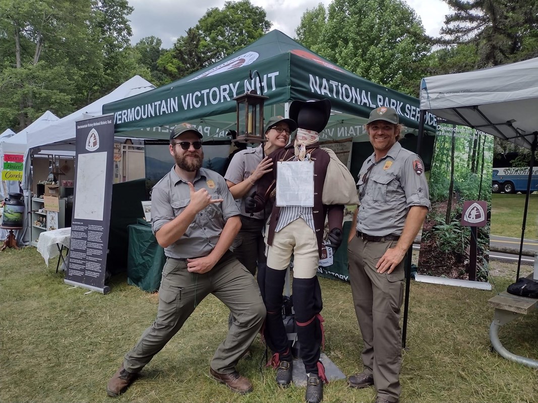 Three people in gray shirts and green pants post together with a mannequin.