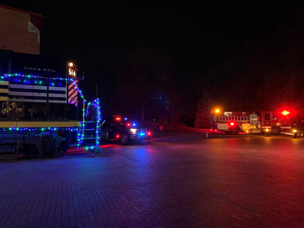 Outside, night time. Train on left, police car in middle, fire truck on right. All lit up
