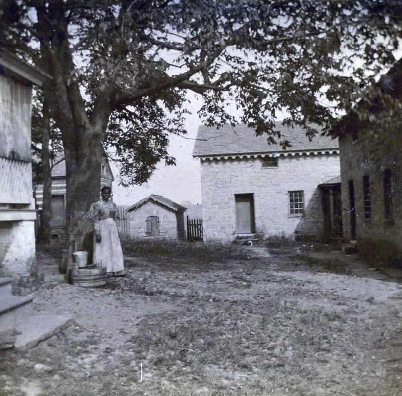 Farmer woman standing in courtyard next to a tree surrounded by slave quarters in 1897