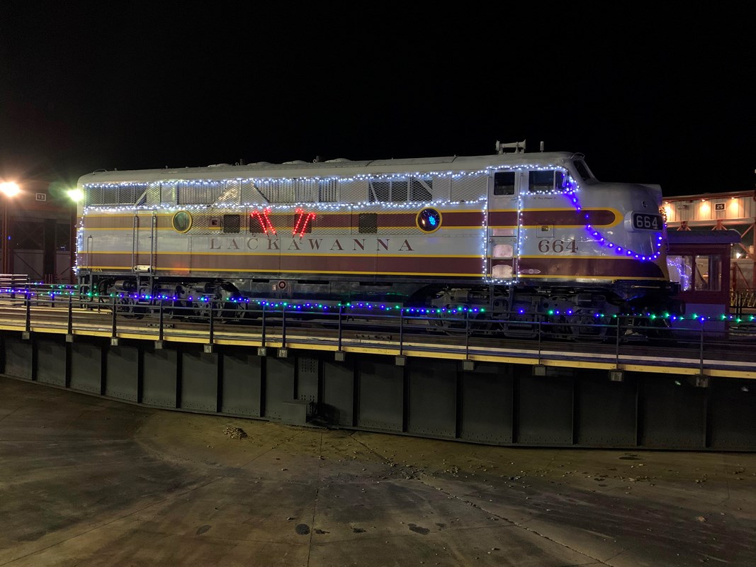 Gray and maroon locomotive decorated in holiday lights, sits on turntable