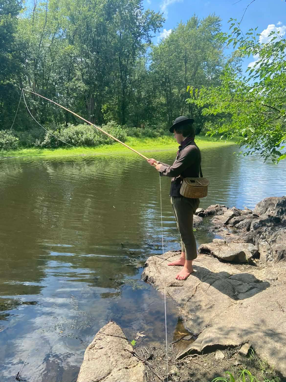 An 18th century man stands at a river fishing. He has a wicker basket on his hip and no shoes on.