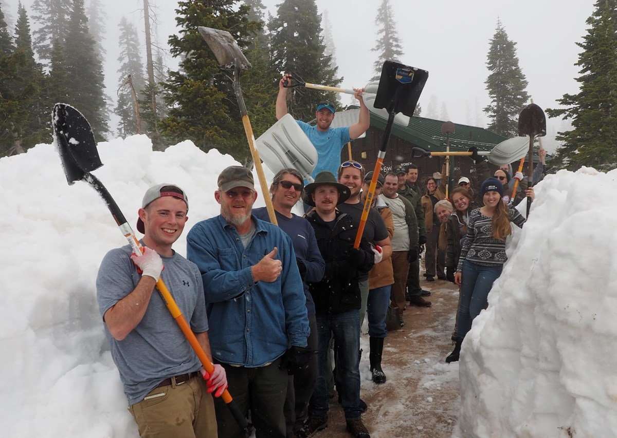 People stand on a cleared path holding shovels with several feet of snow on either side.