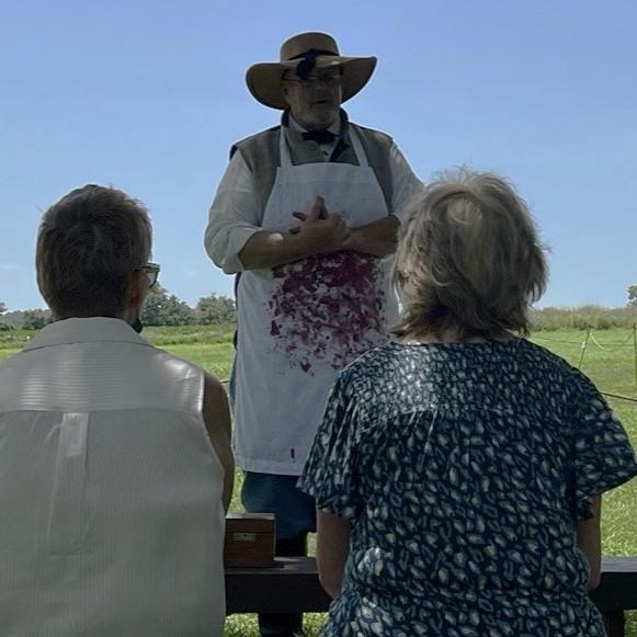A man in living history clothes with imitation blood on the apron speaks with an audience.