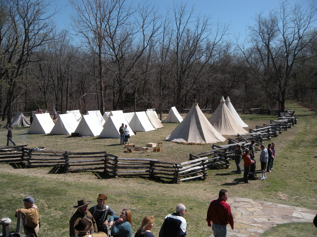 Trees in the background, civil war Sibley Tents and A-frame tents on grassy fenced in area.