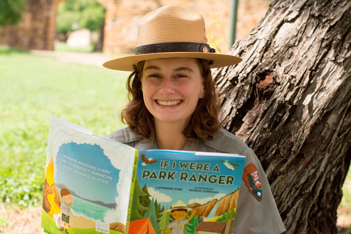 Park Ranger holds book 'If I were a Ranger' while sitting next to a tree at Mission San Jose
