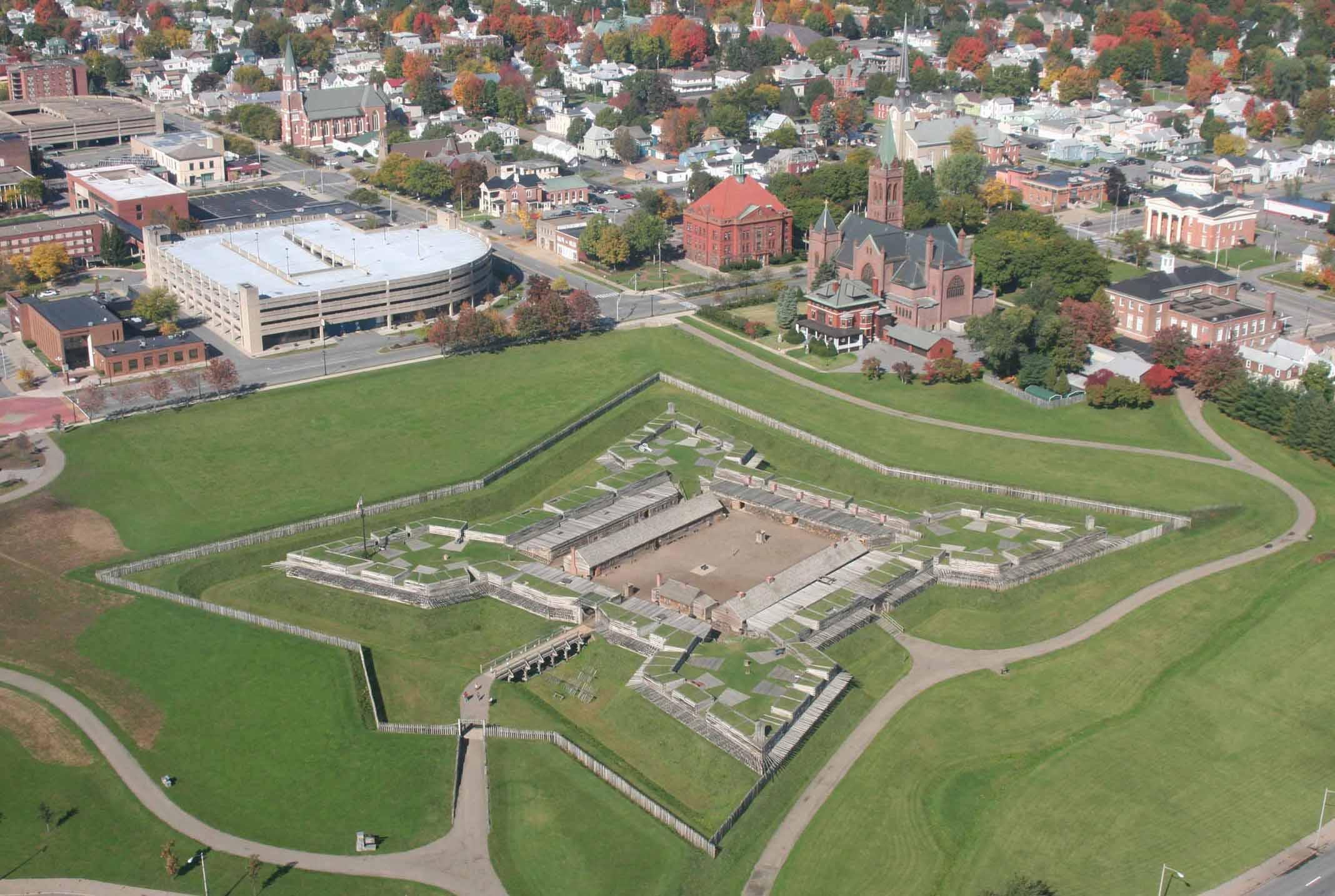 A view of a star-shaped wooden fort in a field of verdant green, from above. A city surrounds it.