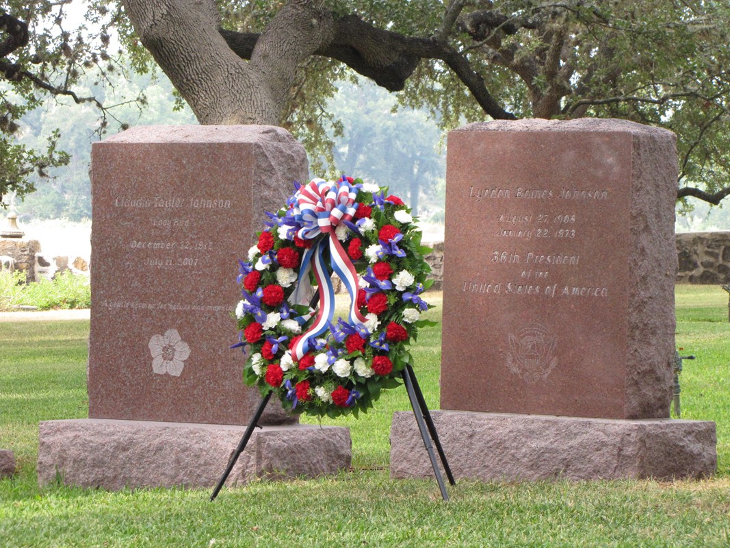 A red, white, and blue wreath stands in front of a pink granite headstone.