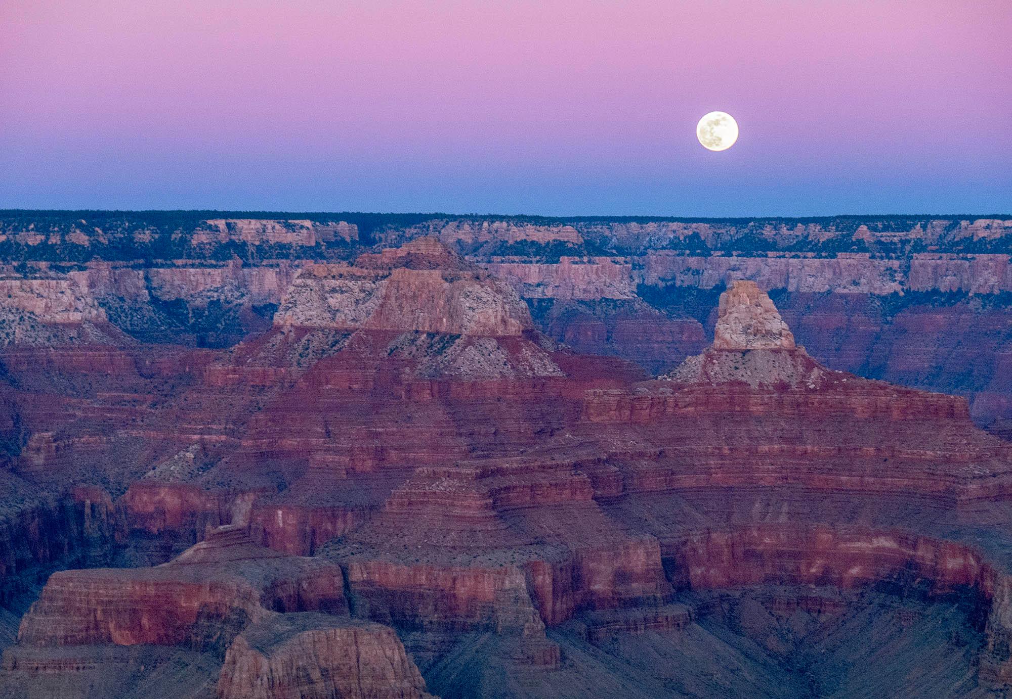 Just after sunset, a full moon is rising above the vermilion peaks and cliffs within Grand Canyon,