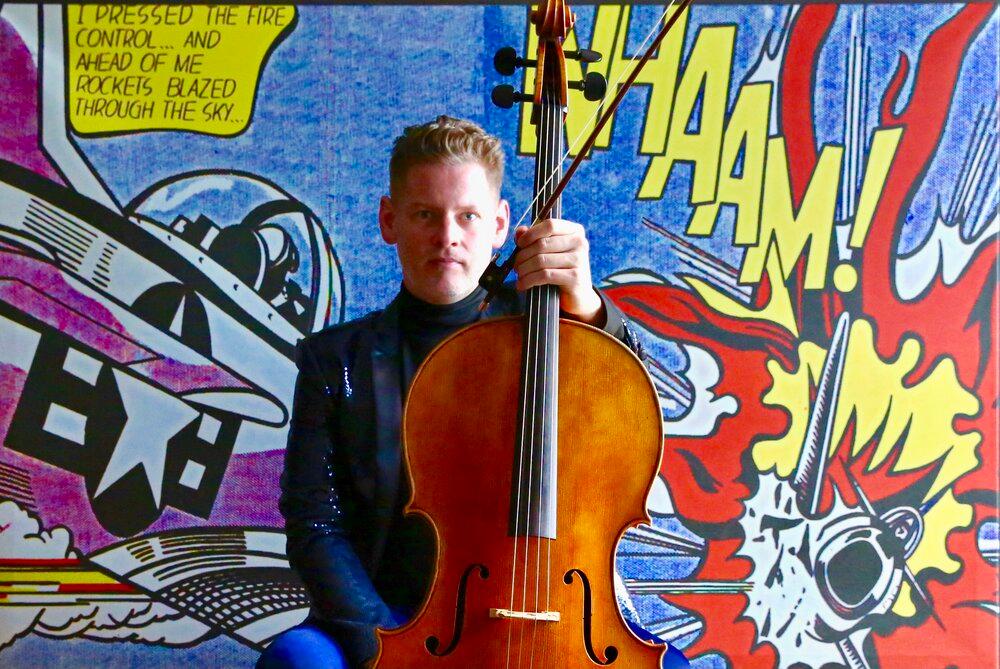 A thirty-something man holding a cello with colorful comic strip characters in the background