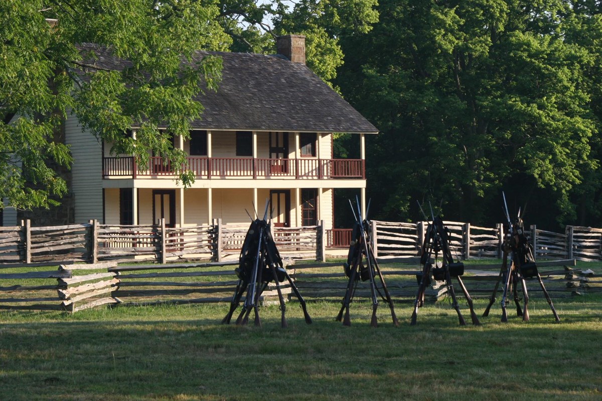 Background of green trees, two-story dog trot house, log fence and stacked muskets in the foreground