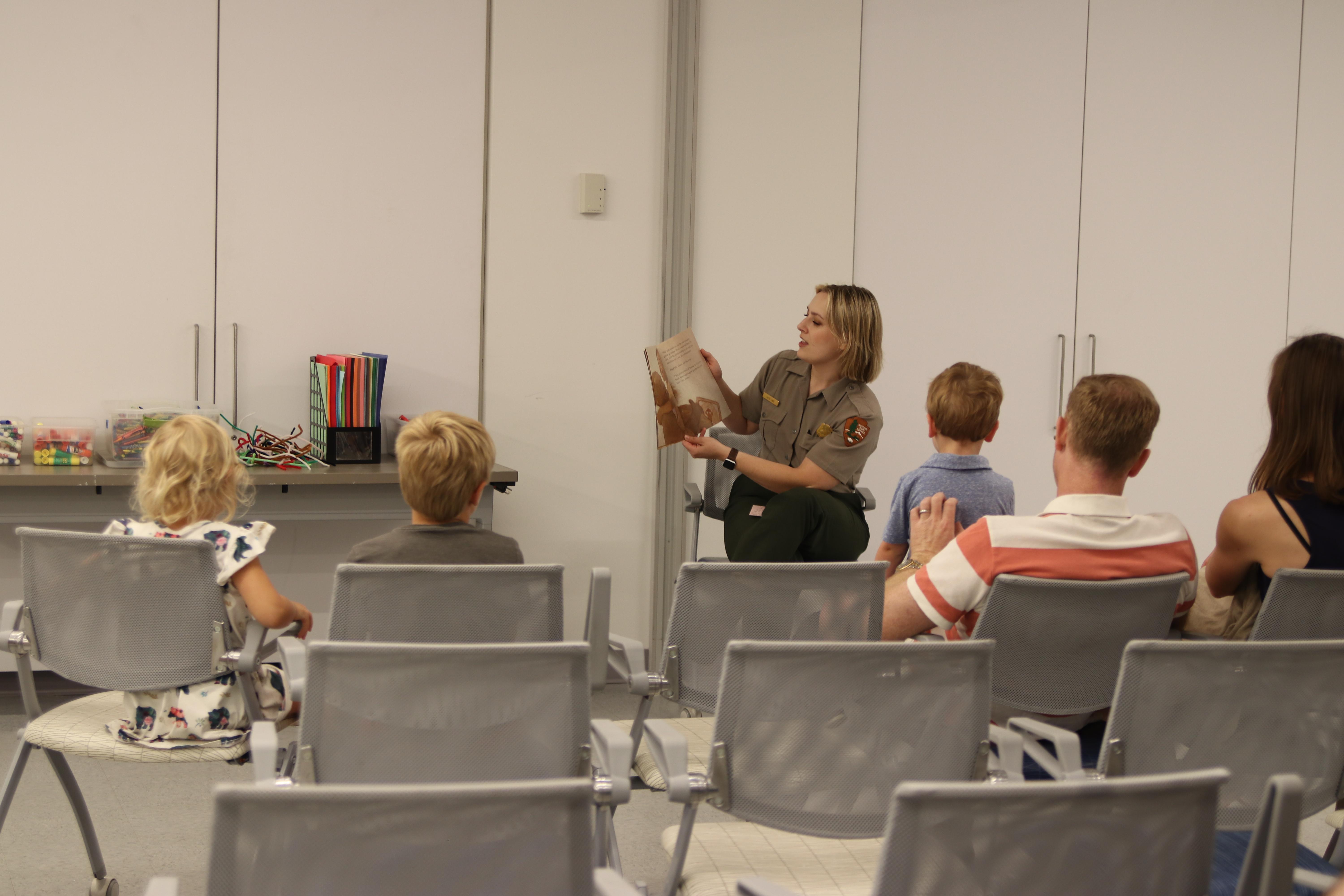 a woman in a ranger uniform holds up a book in front of a small audience of kids and parents