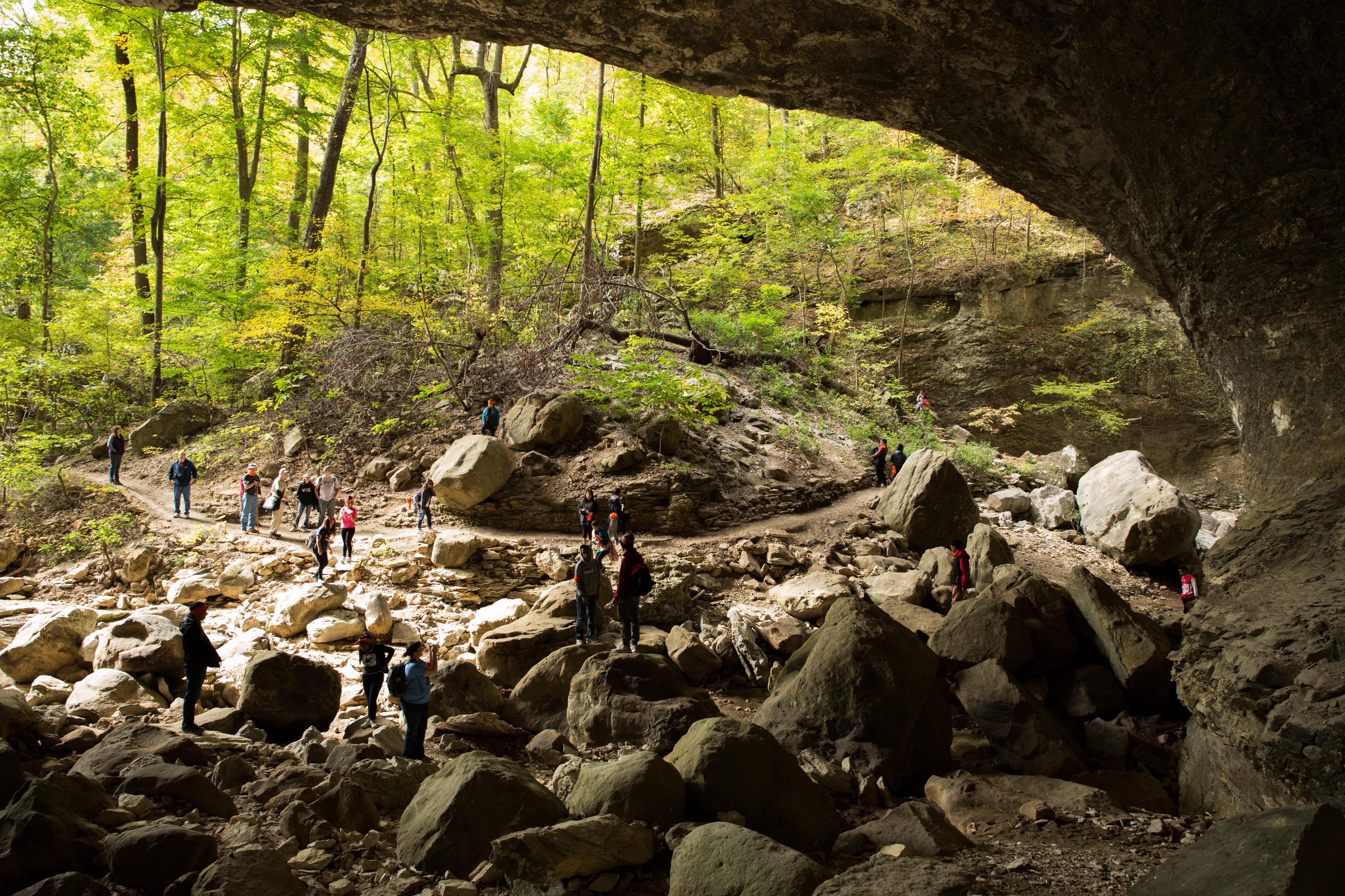 The view looking out from of a bluff shelter towards the woods with people walking on a trail.