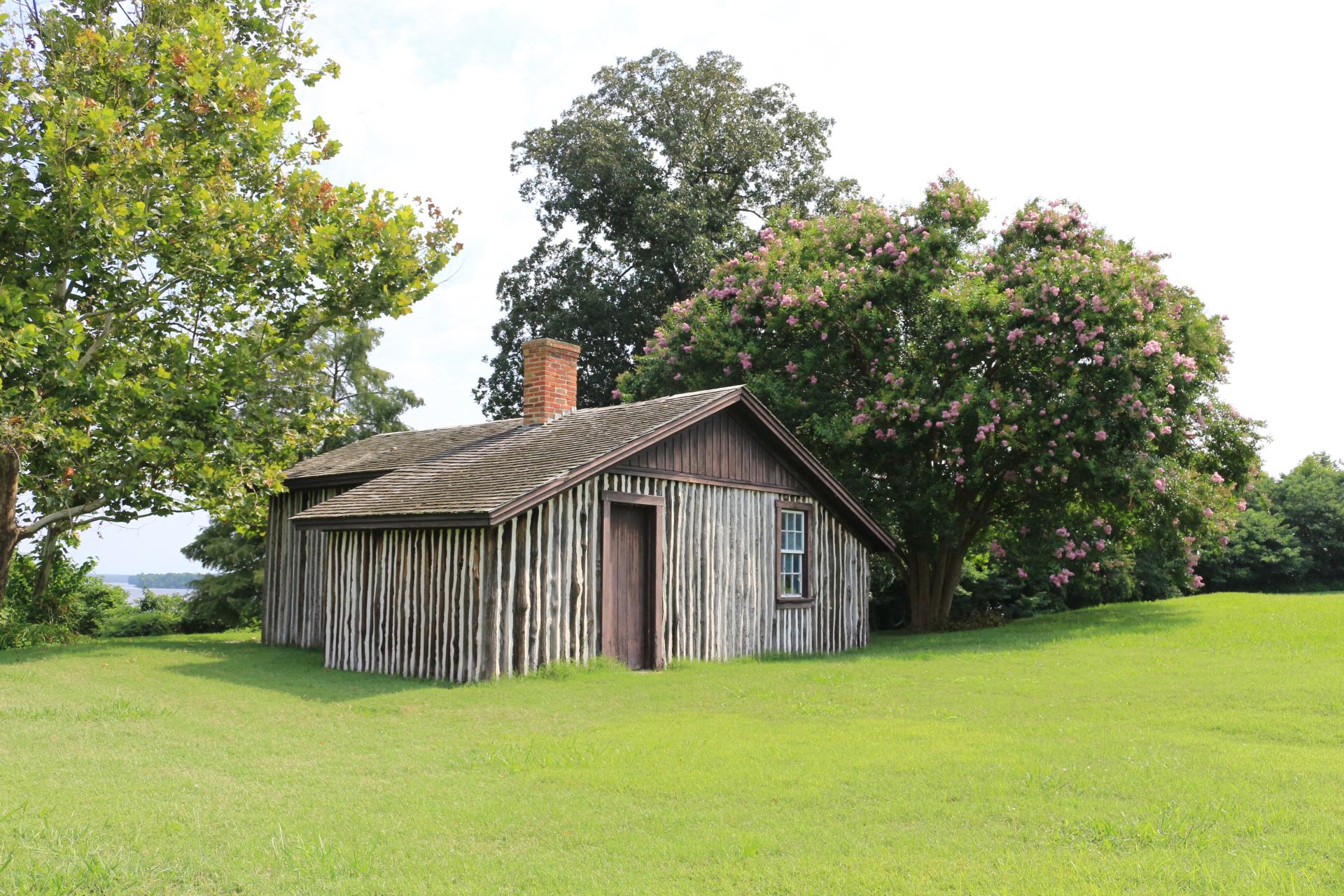 A wooden cabin with a chimney surround by a green lawn and two large trees behind it.