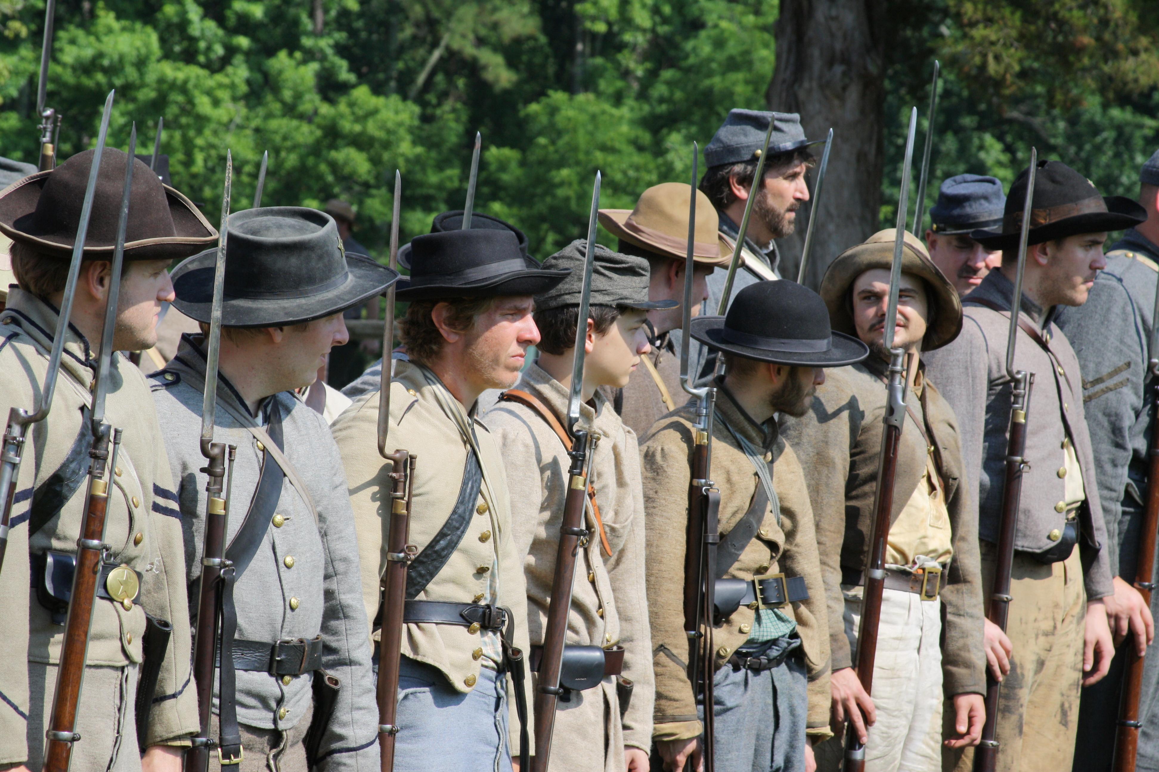 A line of men in gray and butternut Civil War uniforms standing side-by-side at the edge of woods.