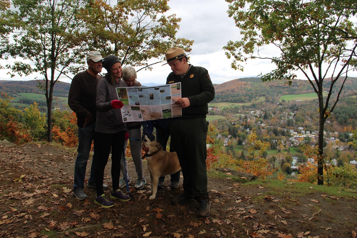 Park Ranger looks at map with 3 visitors and a dog with an autumn overlook behind them