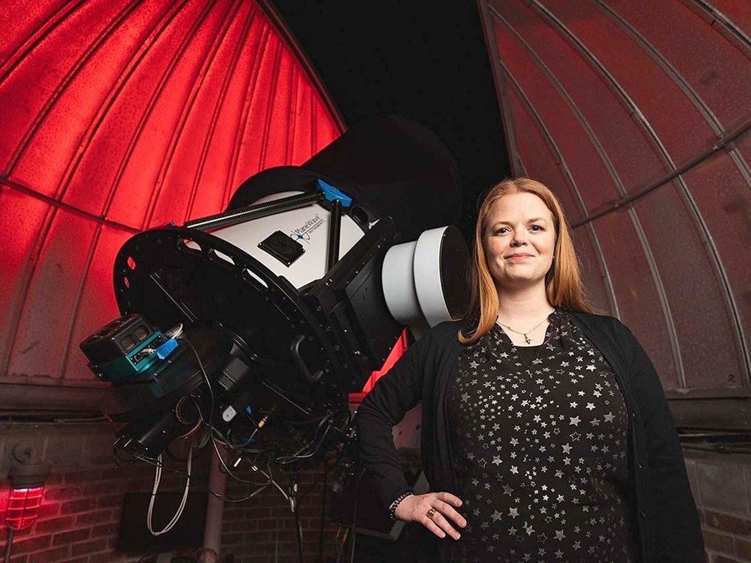 Standing by a telescope, a woman with long, red hair, wears a black dress covered with silver stars