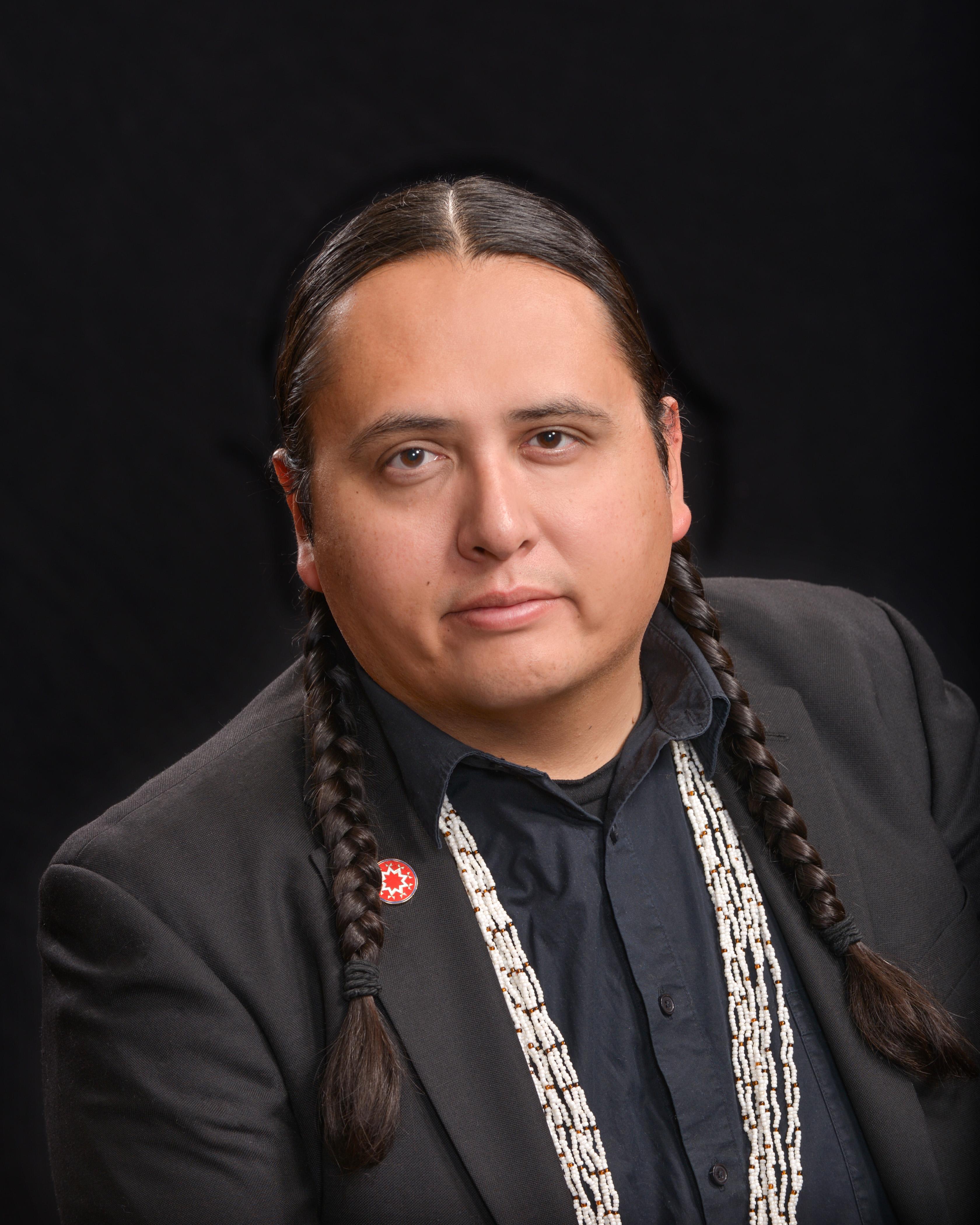 A man wearing a dark suit with braided hair, a beaded necklace and a red lapel pin.