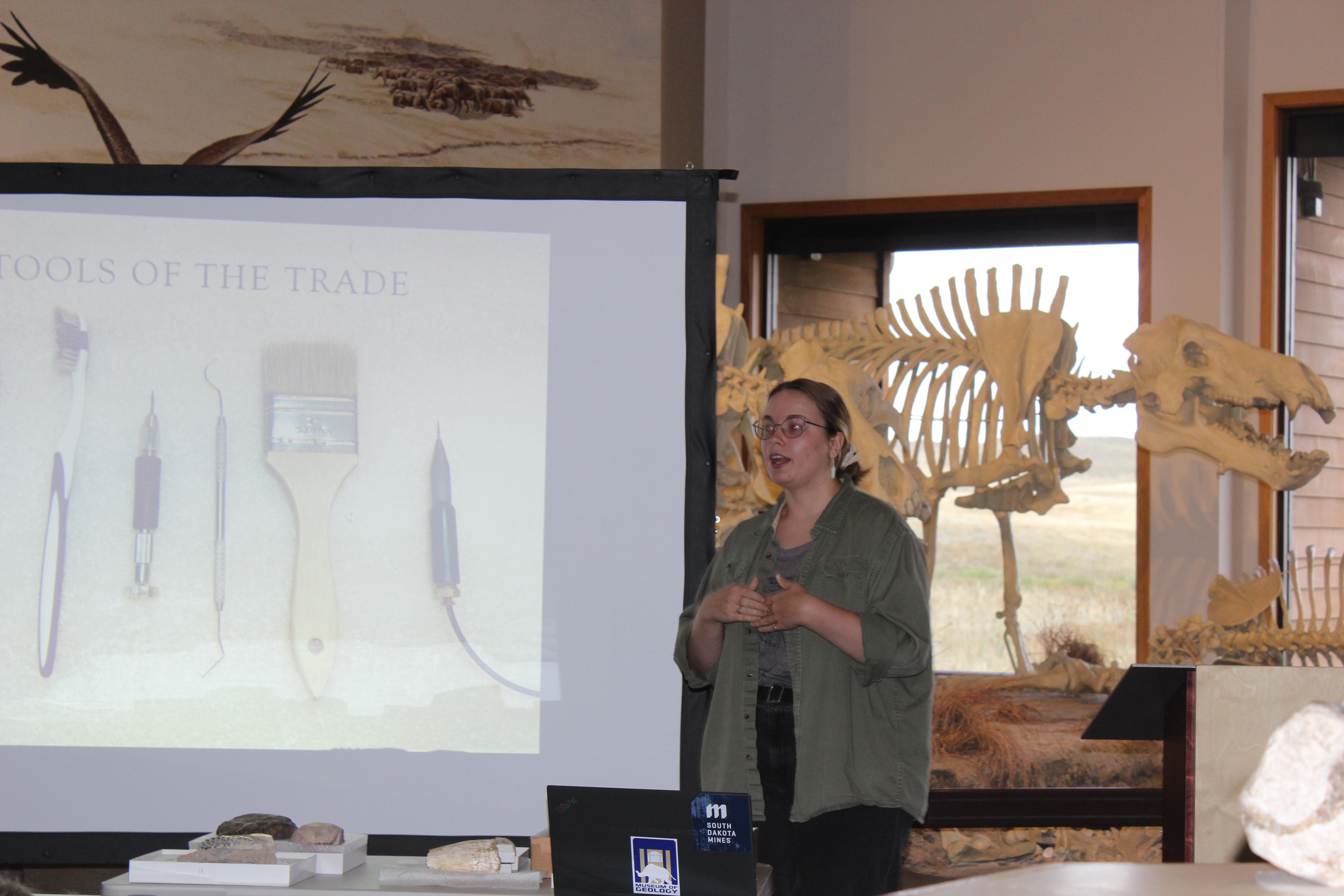 A  woman in plain clothes and glasses stands next to a projector screen showing 5 different tools