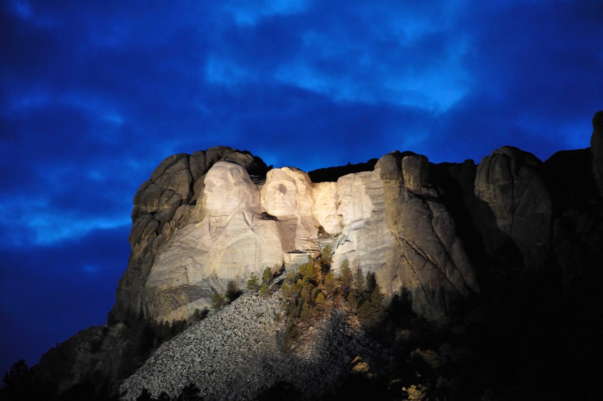 Photo of Mount Rushmore illuminated under a darkening sky.