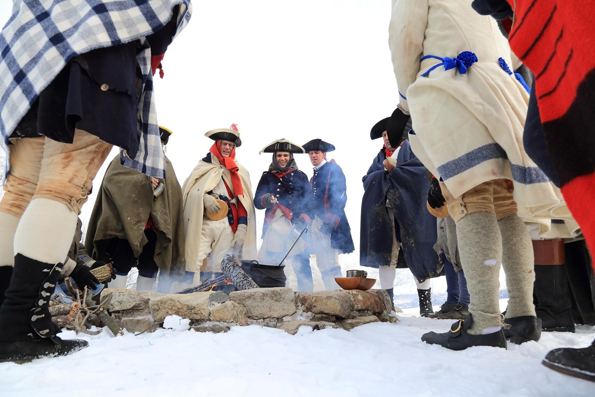 photograph, outdoors, snow, soldiers in eighteenth-century clothing stand around a campfire