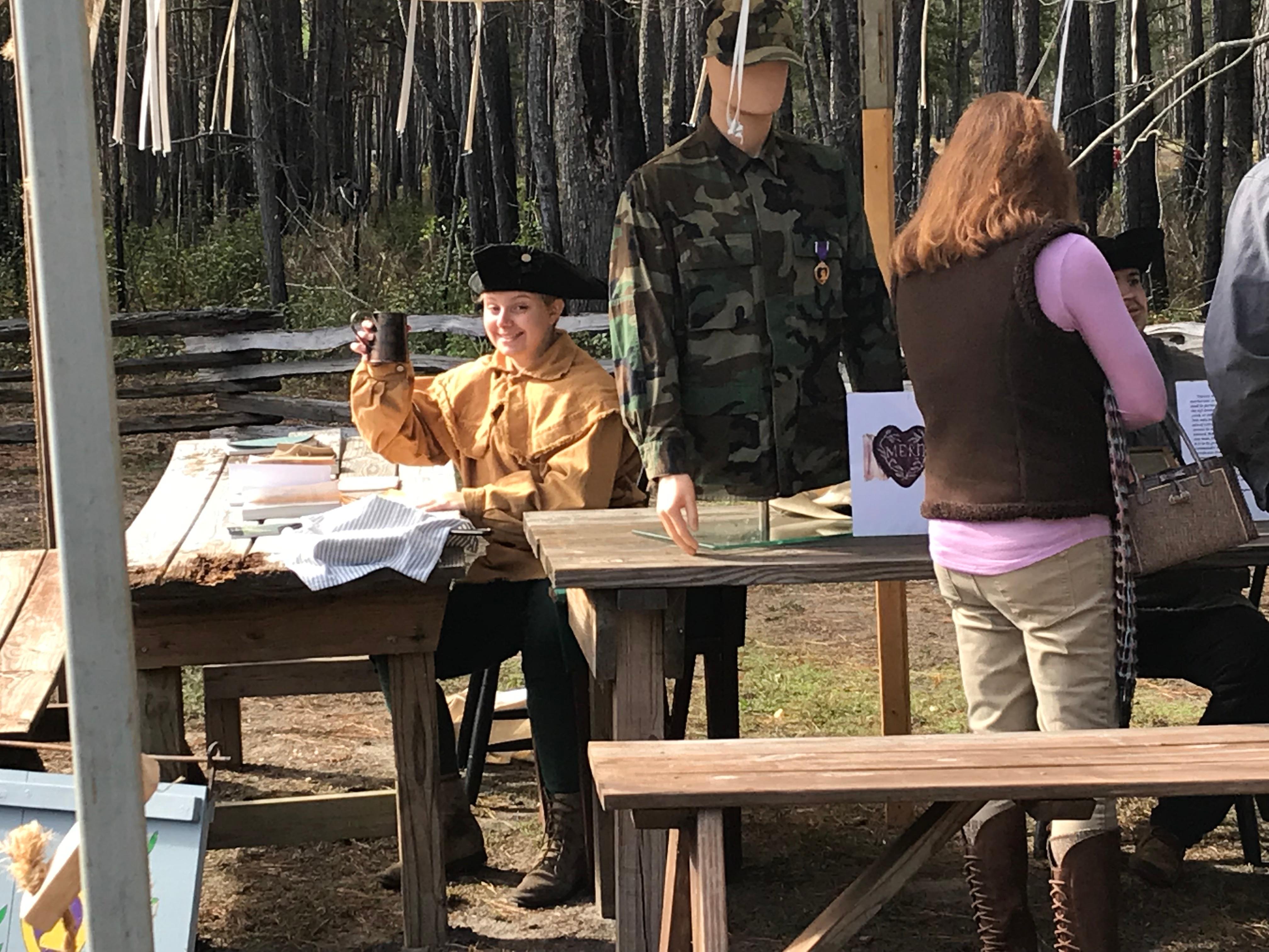 Person wearing colonial clothing sitting at a wooden table with a mannequin on the table.