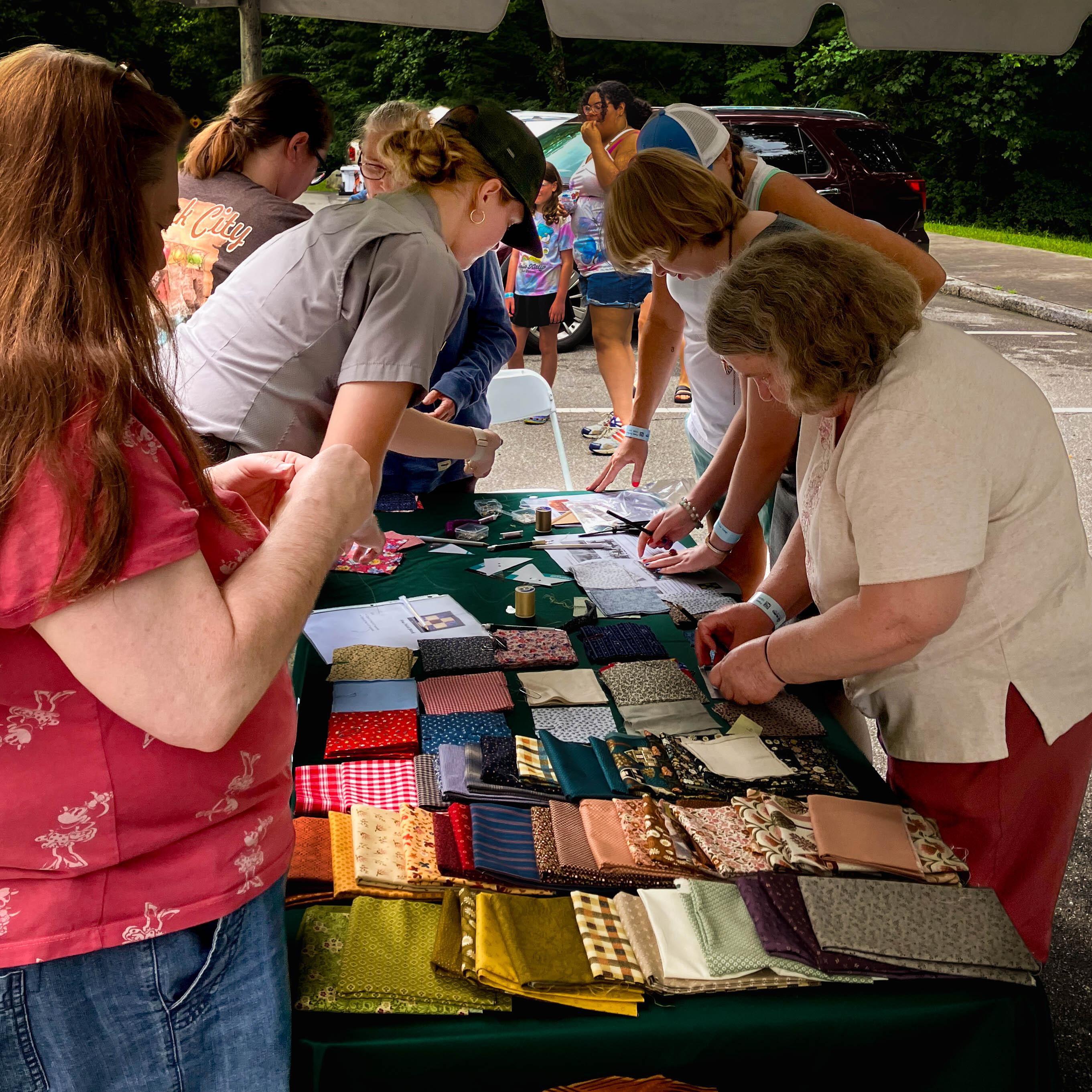 A group of people surrounding a table full or quilting materials and fabrics.