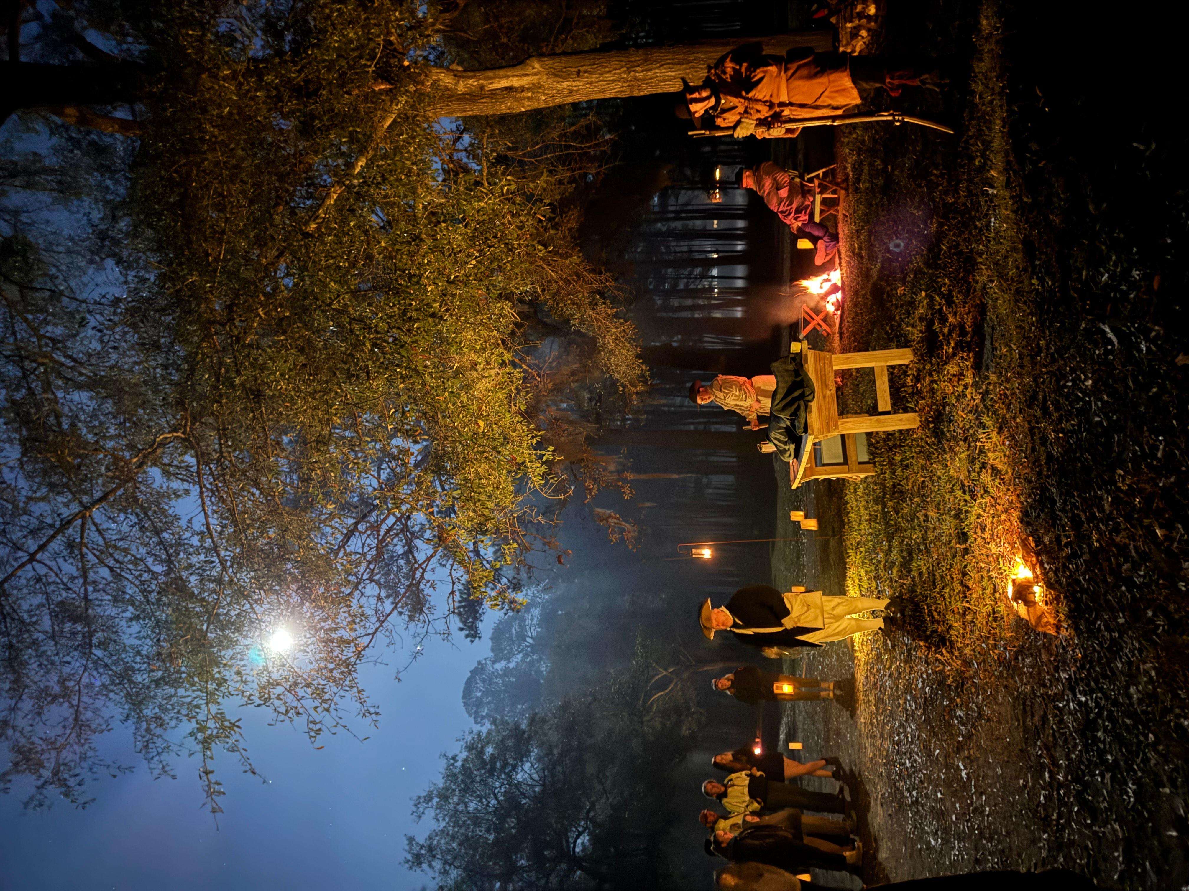 People dressed in colonial clothing outside at night with lanterns and a full moon illuminating them