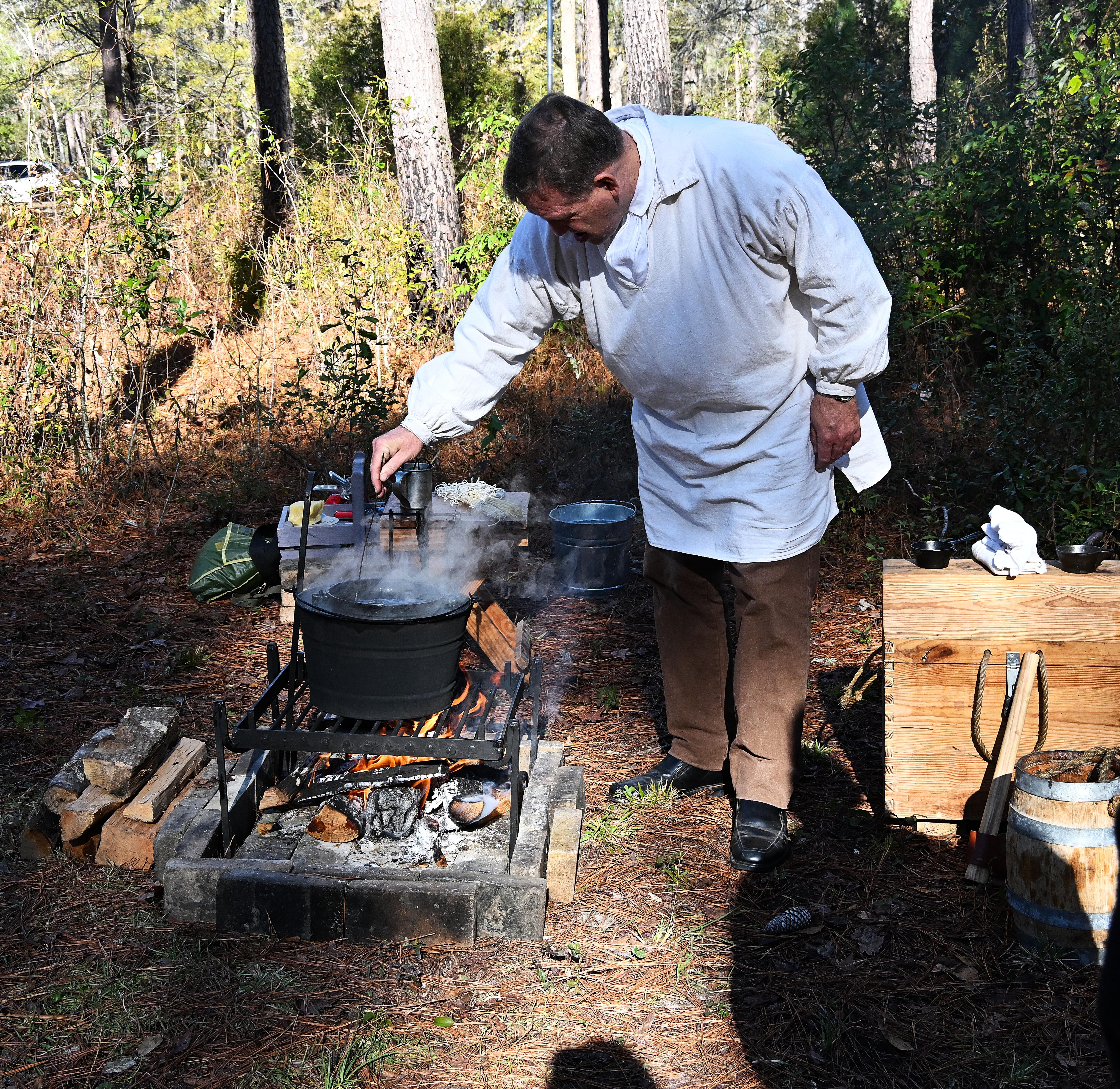 Man in a white smock bending over a campfire dipping a candle