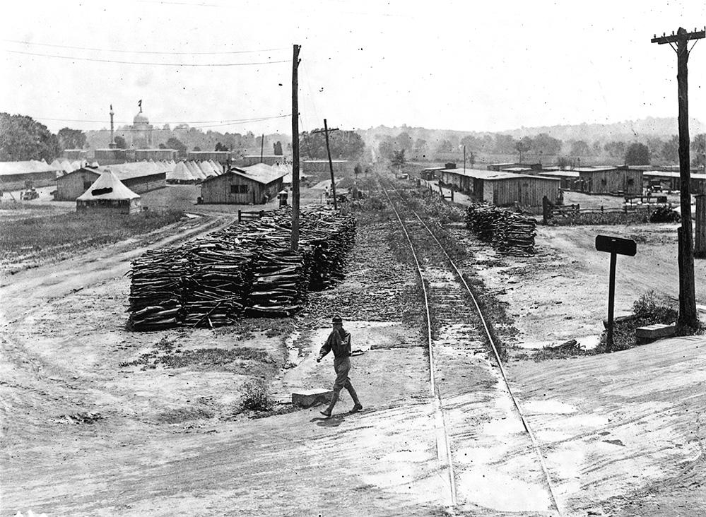 A black and white image showing a soldier walking across a road in front of tents and barracks