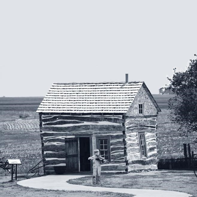 Boy plays fiddle in front of cabin. Image in greyscale.