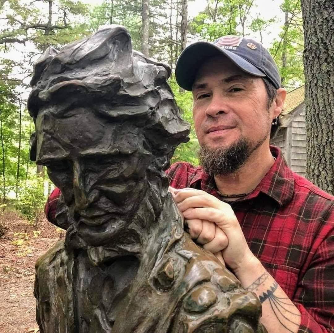 A man in a plaid shirt leans on a bronze monument of Henry David Thoreau