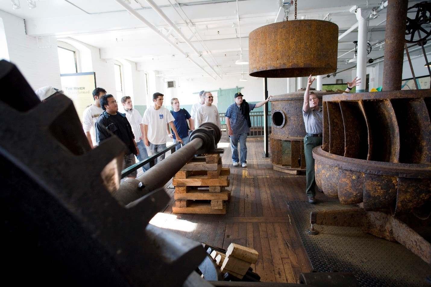 A ranger gestures toward a large metal piece of machinery as a group of visitors look on.