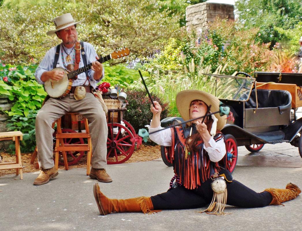 man seated playing banjo. woman doing splits on ground and holding sticks in air
