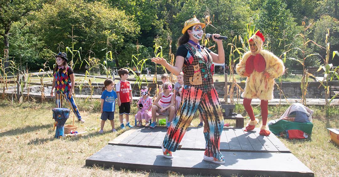 Three performers with face paint and costumes perform a skit in front of corn stalks and children.