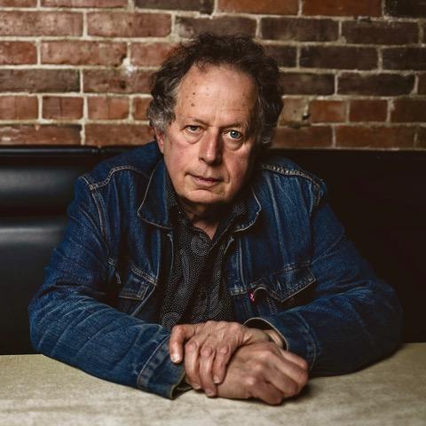 A middle-aged man wearing a denim jacket is seated in front of a brick wall.