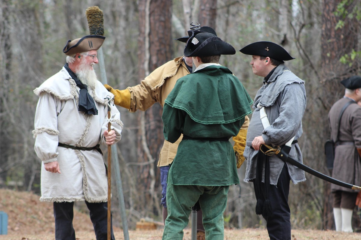 Colonial soldiers dress in civilian attire sanding in a circle talking on an overcast day.