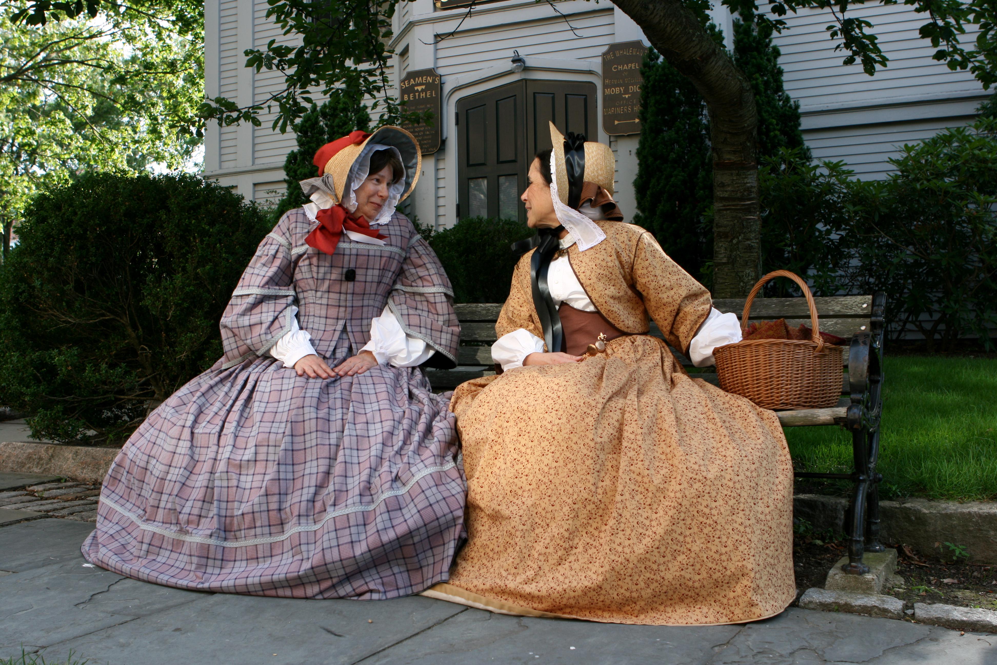 Two people in 1850s-style dresses sit on a bench outside a historic chapel, talking with a basket be