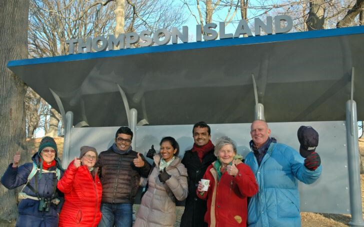 Seven people in winter clothes stand under a sign that reads 