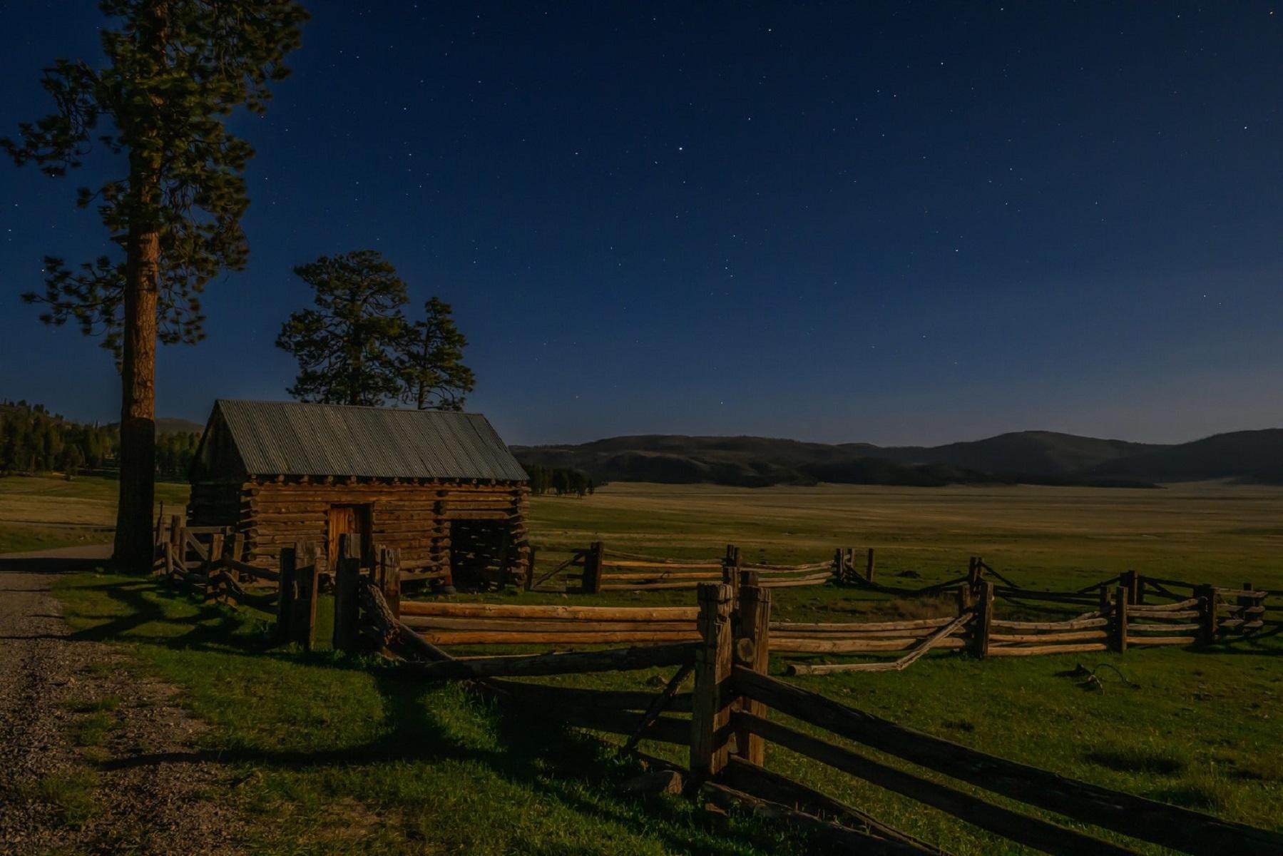 A historic wooden barn and corral in a large valley under the night sky.