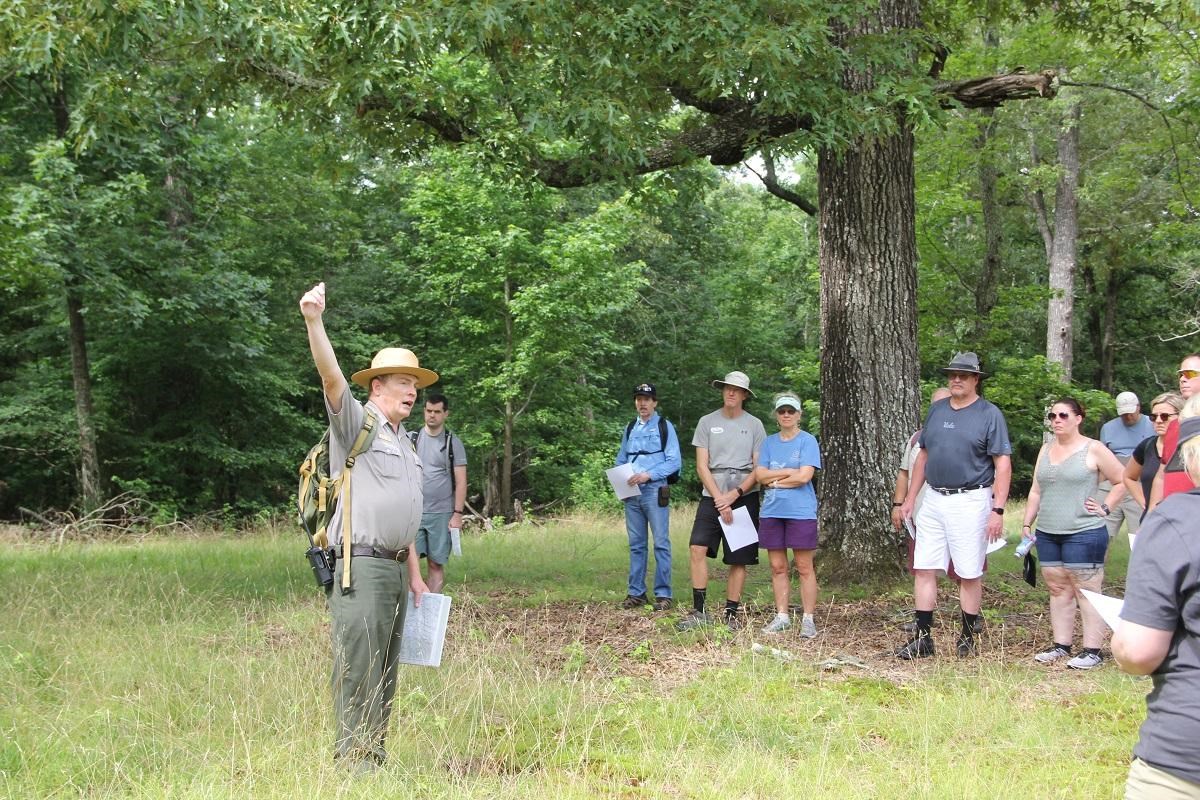 A ranger in green and gray, and wearing a flat hat, points to the sky while leading a hike.