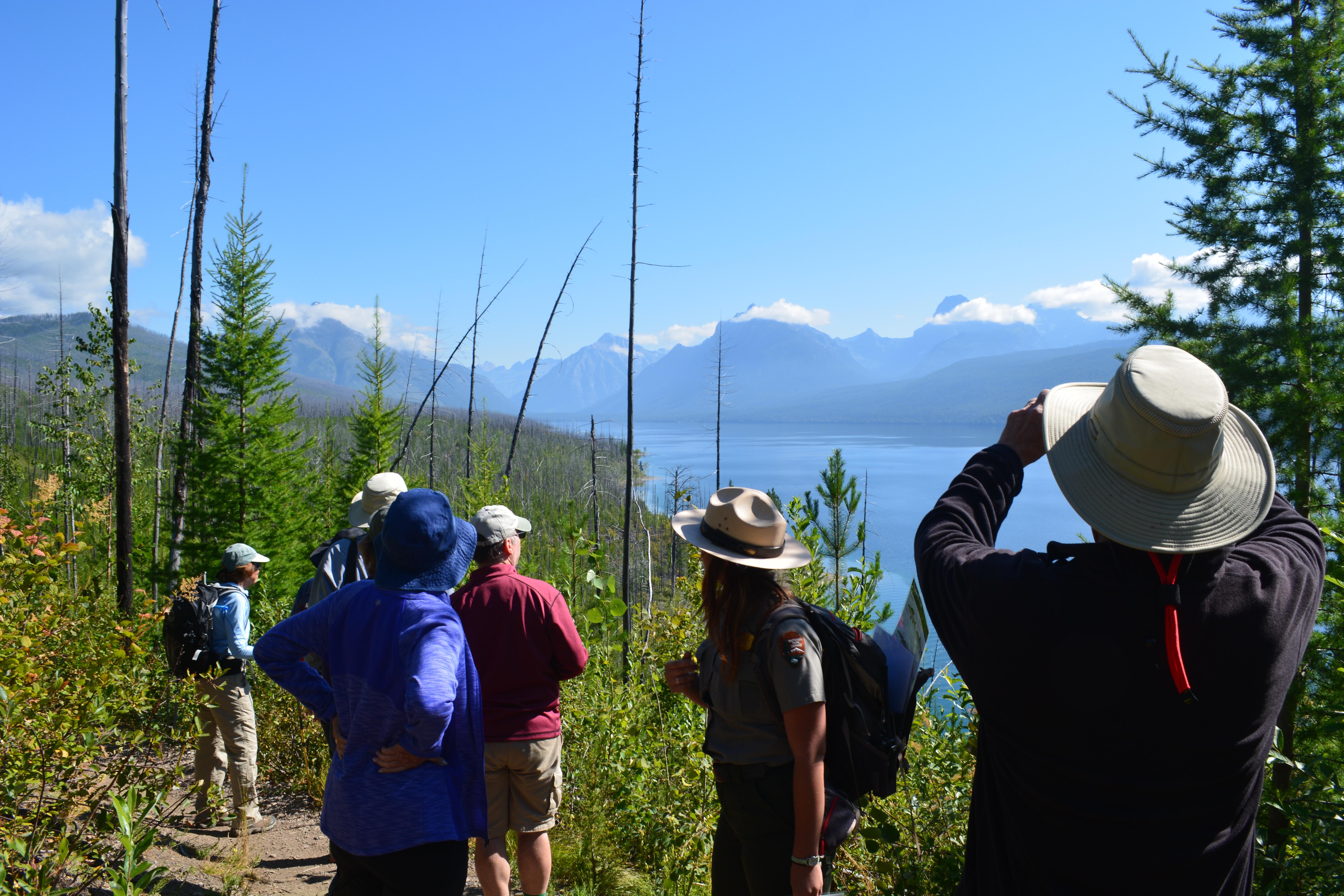 A ranger with visitors in front of mountains and a lake